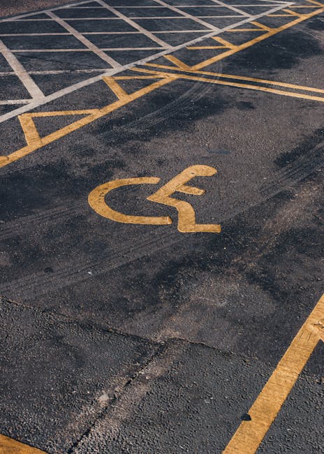 A close-up view of an outdoor parking area surface featuring a yellow painted wheelchair accessibility symbol, placed on dark asphalt with visible texture. Surrounding the symbol are yellow parking boundary lines, some of which are faded or chipped, indicating wear. The surface appears clean but shows signs of weathering and age. The image highlights surface marking visibility, relevant for assessing parking accessibility, which Harringay Cleaners may consider during deep cleaning or maintenance services in commercial or residential parking zones. The lighting suggests daylight, emphasizing the contrast between the asphalt and painted markings. No tools or additional objects are visible in the image. Harringay Cleaners offers cleaning solutions that include surface cleaning and maintenance, ensuring accessible areas remain clear and compliant with regulations.