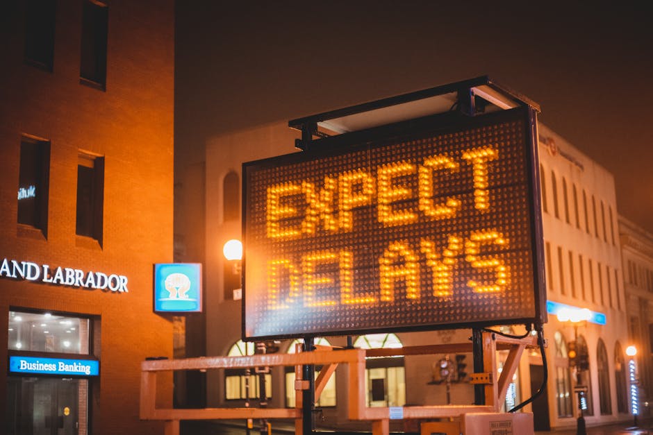 A digital road sign on a city street at night displays the message 'Expect Delays' in bright orange letters. The sign is mounted on a black stand with visible wiring, positioned near a multi-story brick building with illuminated windows. Behind the sign, a blue sign reading 'Land Labrador' and a small illuminated sign for 'Business Banking' can be seen, along with warm street lighting casting a glow on the scene. The street appears quiet with no visible vehicles or pedestrians. This image relates to the topic of access and parking delays for Harringay cleaners, illustrating potential disruptions in urban traffic and parking due to ongoing events or roadworks, emphasizing the importance of advance planning for domestic cleaning appointments with Harringay Cleaners.