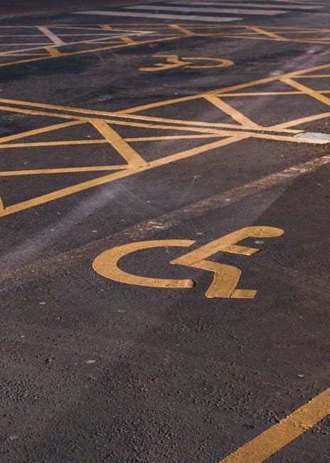 A close-up view of an outdoor parking area surface featuring a yellow painted wheelchair accessibility symbol, placed on dark asphalt with visible texture. Surrounding the symbol are yellow parking boundary lines, some of which are faded or chipped, indicating wear. The surface appears clean but shows signs of weathering and age. The image highlights surface marking visibility, relevant for assessing parking accessibility, which Harringay Cleaners may consider during deep cleaning or maintenance services in commercial or residential parking zones. The lighting suggests daylight, emphasizing the contrast between the asphalt and painted markings. No tools or additional objects are visible in the image. Harringay Cleaners offers cleaning solutions that include surface cleaning and maintenance, ensuring accessible areas remain clear and compliant with regulations.