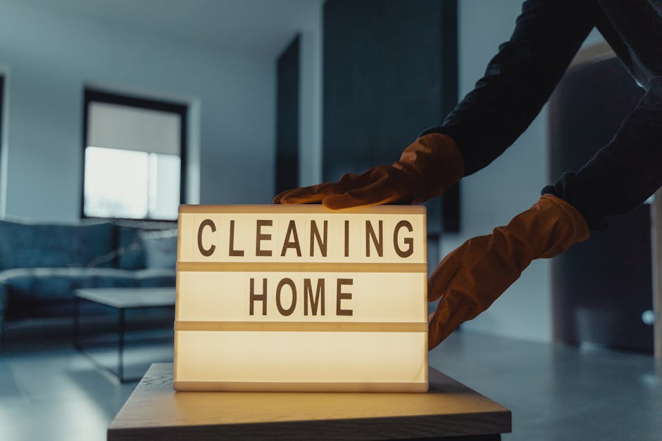 A person wearing light brown cleaning gloves is adjusting a small illuminated sign on a wooden surface. The sign displays the words 'CLEANING HOME' in bold, black letters on white background panels. In the background, a modern living room with a grey sofa and a coffee table is visible, illuminated by natural daylight coming through a large window. The room has a clean and tidy appearance, emphasizing a well-maintained and hygienic environment, consistent with domestic cleaning services offered by Harringay Cleaners. The overall scene suggests a focus on surface cleaning and home sanitisation, highlighting the professional cleaning process in a residential setting.