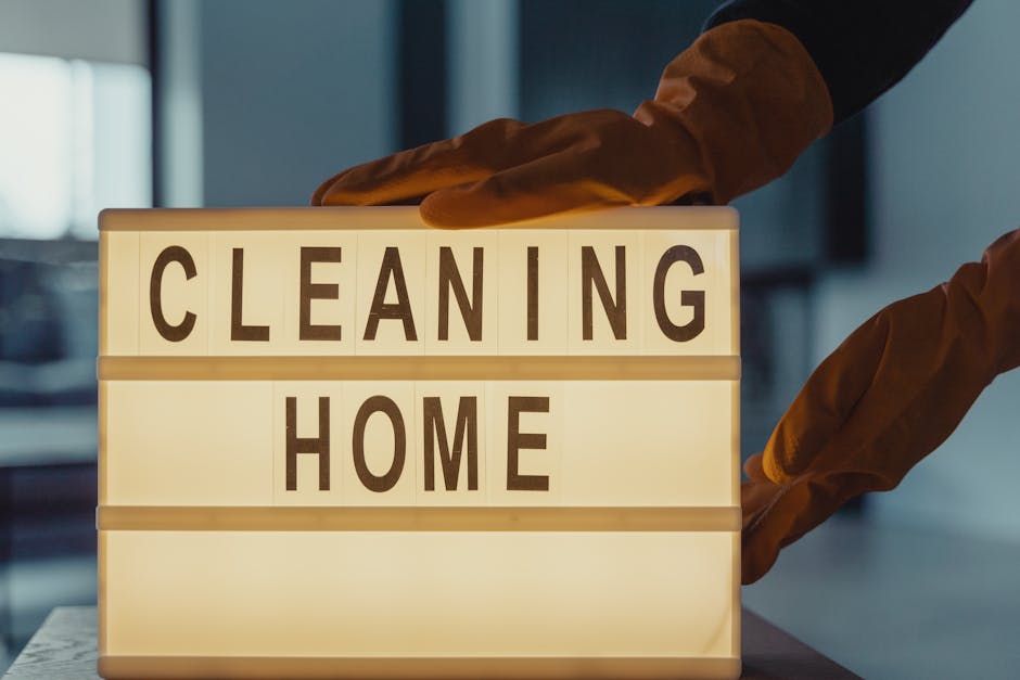 A person wearing orange cleaning gloves is holding a lightbox display with the words 'CLEANING HOME' in black capital letters on illuminated white background panels. The background shows a blurred indoor setting with natural light entering through a window, suggesting a clean and tidy space. This image emphasizes surface cleaning and hygiene associated with domestic cleaning services promoted by Harringay Cleaners, as discussed on the page 'Avoid hidden cleaning charges in Harringay quotes' on harringaycleaners.org.uk.