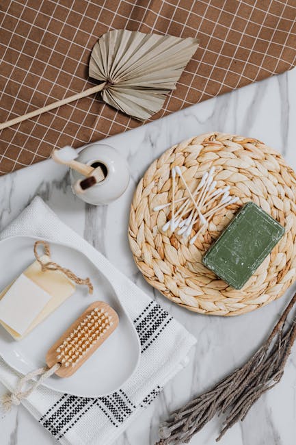 A close-up of a bathroom countertop featuring a woven circular placemat holding a green bar of soap and cotton swabs, with a ceramic soap dispenser nearby. The surface appears clean and well-maintained. Beside the placemat, there is a small, dried leaf-shaped decorative object and a wooden massage brush with a rope handle resting on a white towel with black embroidery. The countertop itself is made of white marble with subtle gray veining, and the background includes a brown tiled wall providing a warm, neutral setting. The arrangement reflects attention to hygiene and surface cleanliness, representative of domestic cleaning practices highlighted by Harringay Cleaners on their website.