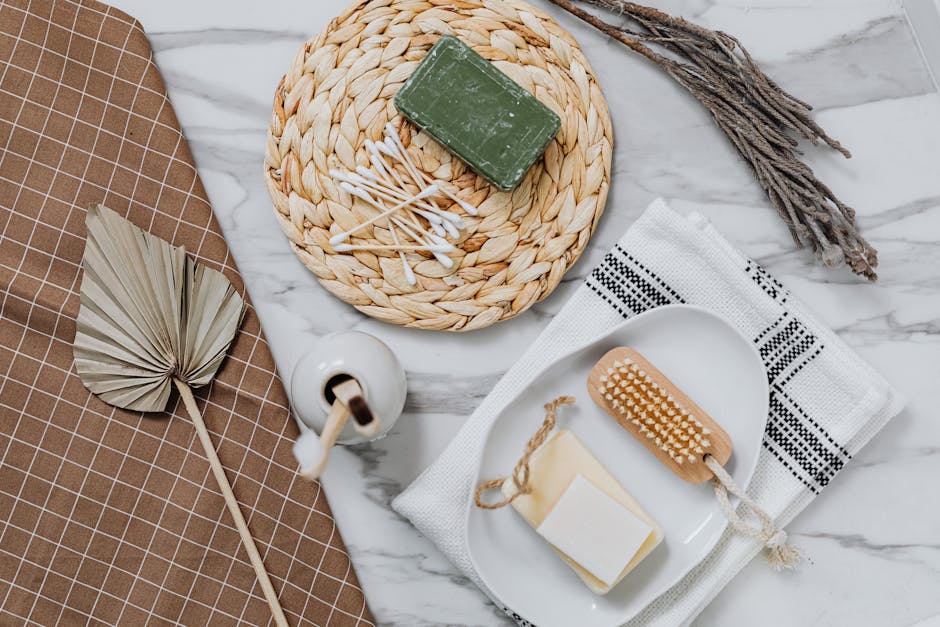 A close-up of a bathroom countertop featuring a woven circular placemat holding a green bar of soap and cotton swabs, with a ceramic soap dispenser nearby. The surface appears clean and well-maintained. Beside the placemat, there is a small, dried leaf-shaped decorative object and a wooden massage brush with a rope handle resting on a white towel with black embroidery. The countertop itself is made of white marble with subtle gray veining, and the background includes a brown tiled wall providing a warm, neutral setting. The arrangement reflects attention to hygiene and surface cleanliness, representative of domestic cleaning practices highlighted by Harringay Cleaners on their website.