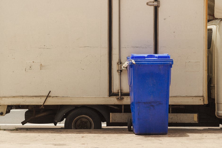 A blue recycling bin with a lid is positioned in front of a white delivery van, partially blocking the lower part of the vehicle. The surface of the van appears to be made of metal and shows signs of 