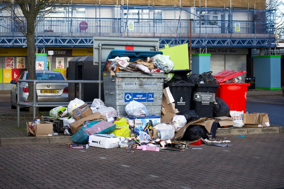 A pile of overflowing mixed paper, cardboard, and plastic waste bags, along with boxes and miscellaneous trash, dumped next to a large grey recycling bin with blue and white labels indicating mixed paper and card recycling. Beside it, there are black and red waste bins, some with lids open revealing additional rubbish. The scene is set on a paved sidewalk in front of a commercial building with storefronts, visible signage, and a metal railing, under natural daylight with a tree on the left. The image illustrates improper waste disposal outside a commercial area, emphasizing the importance of following waste management rules in Harringay, related to the services provided by Harringay Cleaners.