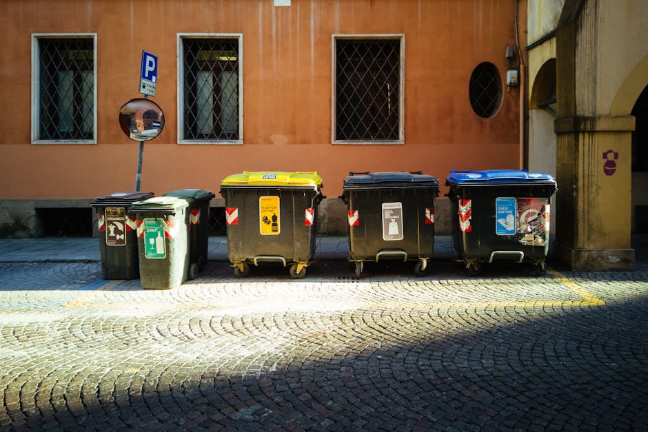 A row of four large outdoor waste bins positioned along a cobblestone sidewalk in front of a building with a salmon-colored wall. The bins have different colored lids—green, yellow, black, and blue—and feature recycling and waste symbols. The area around the bins appears clean and well-maintained, with sunlight casting shadows on the cobblestones. Behind the bins, there is a pole with a parking sign and windows with metal grilles on the building's wall. The scene reflects ongoing waste disposal practices as recommended by Haringey Council, with Harringay Cleaners emphasizing proper surface cleaning and waste management.