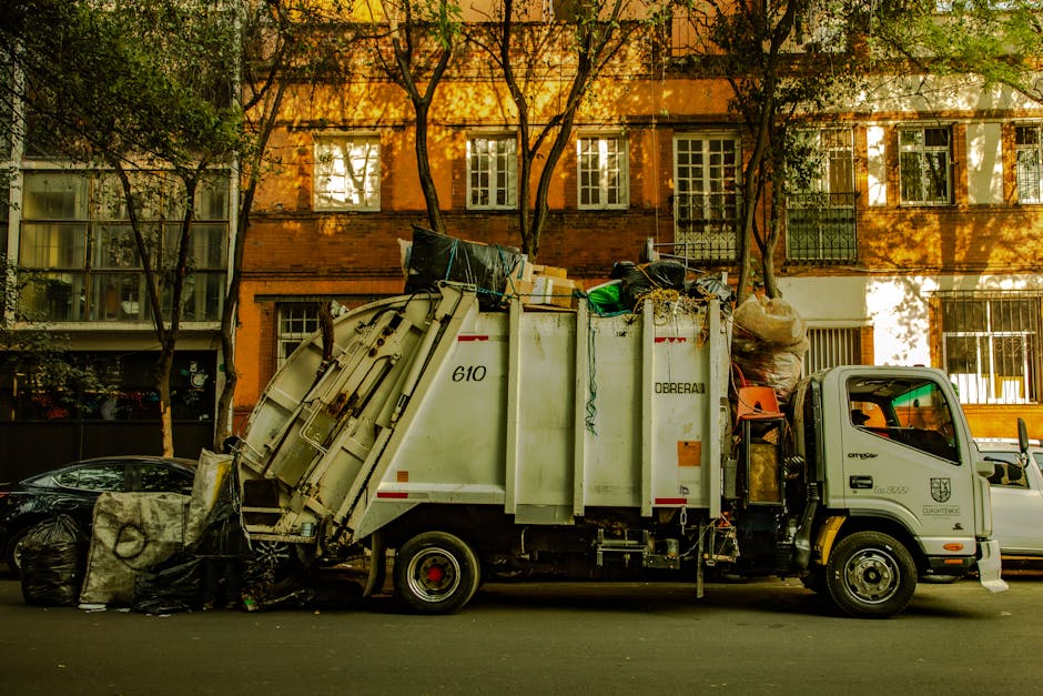 A pile of overflowing mixed paper, cardboard, and plastic waste bags, along with boxes and miscellaneous trash, dumped next to a large grey recycling bin with blue and white labels indicating mixed paper and card recycling. Beside it, there are black and red waste bins, some with lids open revealing additional rubbish. The scene is set on a paved sidewalk in front of a commercial building with storefronts, visible signage, and a metal railing, under natural daylight with a tree on the left. The image illustrates improper waste disposal outside a commercial area, emphasizing the importance of following waste management rules in Harringay, related to the services provided by Harringay Cleaners.