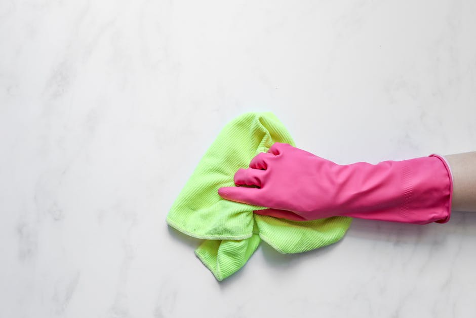 A person wearing a bright pink rubber cleaning glove is holding a lime green microfiber cloth, wiping a smooth, white marble surface with subtle grey veining. The surface appears clean and polished, reflecting soft natural light with minimal dust or smudges. The background is plain, emphasizing the surface and cleaning tools used for surface cleaning and maintenance. This image illustrates domestic cleaning practices, highlighting the use of effective materials and tools, consistent with the services offered by Harringay Cleaners, especially for end of tenancy cleaning and sanitisation procedures.