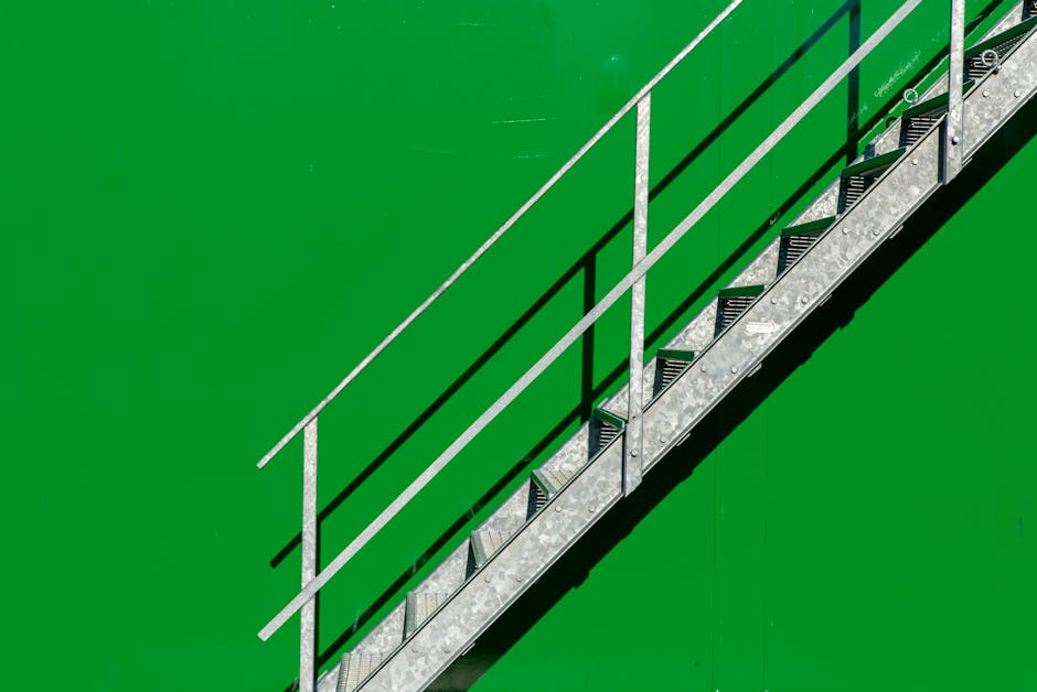 A high-angle view of a metal staircase with black anti-slip treads, set against a vibrant green surface, casting clear shadows. The staircase has silver handrails on both sides, with a smooth, clean finish, and is illuminated by natural light highlighting its metallic shine. This staircase is part of a residential or commercial space, emphasizing surface cleaning and maintenance. Harringay Cleaners specializes in deep cleaning and sanitisation, ensuring such surfaces remain spotless and safe, especially in areas with tight stair access, as referenced in the Harringay Ladder flat cleaning tips for tight stair access. The image showcases the importance of proper surface cleaning and upkeep of metallic and painted structures to achieve hygienic and visually appealing environments.