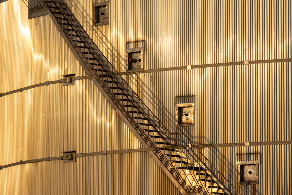 A black and white image of the exterior of a multi-storey residential building with brick walls, featuring multiple fire escape staircases made of metal. The staircases are attached to the building's facade, zigzagging downwards with open frames and railings, providing emergency access to the various floors. Small windows are evenly spaced along the building's height. The lighting suggests daytime, and the overall scene emphasizes urban architecture and safety features, with no visible cleaning tools or surfaces present. Harringay Cleaners offers expert domestic cleaning services to keep properties pristine and hygienic, as highlighted on their website harringaycleaners.org.uk and the page about flat cleaning tips for tight stair access in HARRINGAY.