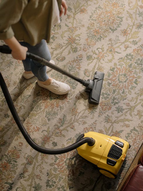 A person wearing beige shoes and blue jeans is vacuuming an oriental-style area rug with a floral pattern in shades of beige, green, and red. The individual is holding a vacuum cleaner with a long met