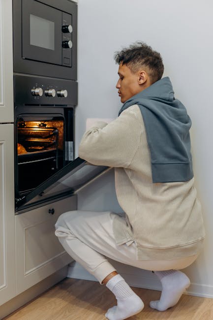 A young man with short black hair, wearing a beige hoodie with a grey sweatshirt draped over his shoulders, beige joggers, and white socks, kneeling on the floor while opening the door of a built-in oven in a modern kitchen. The oven is stainless steel with a digital display and dials, and the interior appears clean and illuminated. The kitchen features white cabinetry and a light wooden floor, with bright, neutral lighting highlighting the surface cleanliness. Harringay Cleaners provides professional domestic cleaning services, including deep oven cleaning to ensure hygiene and optimal performance, as depicted in this scene of routine kitchen maintenance.