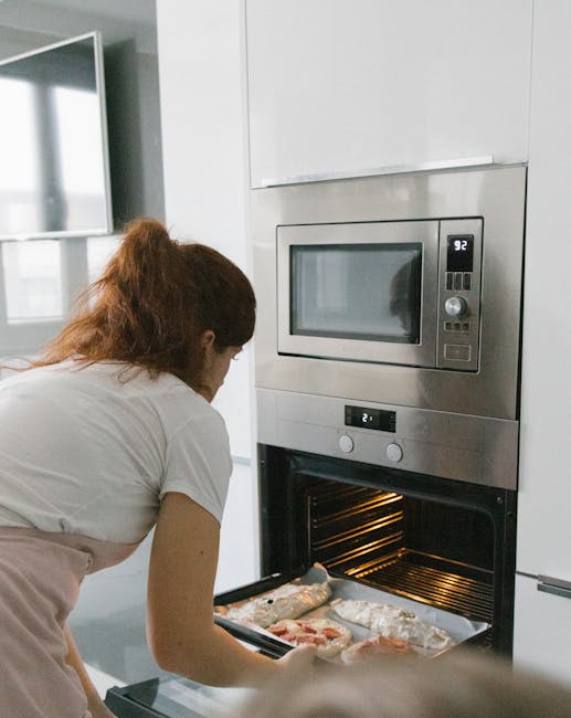 A woman with long, reddish-brown hair tied back is placing baking trays with foil into a modern built-in oven in a kitchen with white cabinetry. The oven, featuring a stainless steel finish, has a digital control panel and a microwave above it. The interior of the oven is illuminated, revealing multiple wire racks, and the surrounding surfaces are clean and free of grease. Natural light from a nearby window reflects softly off the smooth, white wall behind her. This scene exemplifies professional surface cleaning and oven sanitisation services as promoted by Harringay Cleaners for same-day domestic cleaning and oven cleaning in Turnpike Lane, Haringay.
