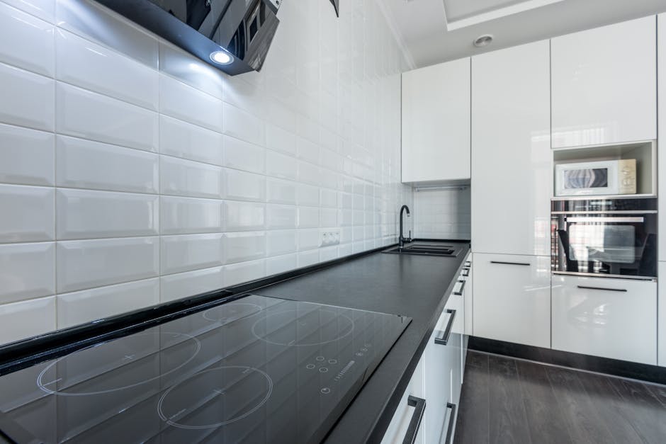 A young man with short black hair, wearing a beige hoodie with a grey sweatshirt draped over his shoulders, beige joggers, and white socks, kneeling on the floor while opening the door of a built-in oven in a modern kitchen. The oven is stainless steel with a digital display and dials, and the interior appears clean and illuminated. The kitchen features white cabinetry and a light wooden floor, with bright, neutral lighting highlighting the surface cleanliness. Harringay Cleaners provides professional domestic cleaning services, including deep oven cleaning to ensure hygiene and optimal performance, as depicted in this scene of routine kitchen maintenance.