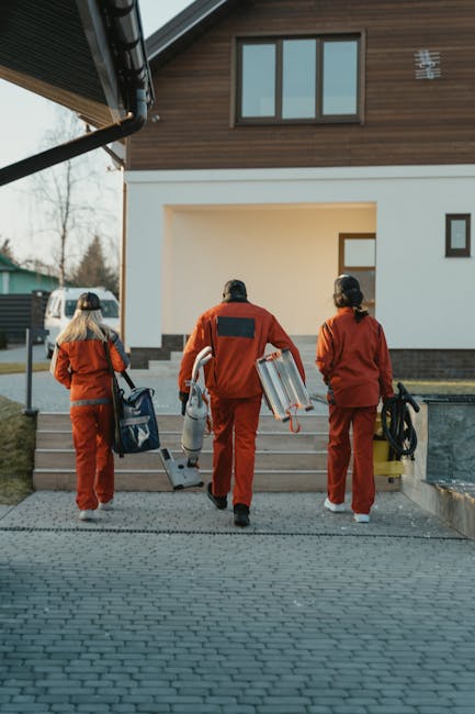 Three professional cleaners dressed in orange uniforms with black accents, carrying cleaning equipment such as vacuum cleaners, spray bottles, and buckets, walking towards the entrance of a modern residential property with a wooden and white facade. The scene takes place outdoors on a paved pathway leading to steps, with natural daylight illuminating the area. The house features large windows and a well-maintained exterior, indicating a residential setting in Harringay. Harringay Cleaners provides urgent flood cleaning and drying services, ensuring surfaces like floors and walls are thoroughly cleaned and dried from water damage, with attention to hygiene and surface sanitation applicable to domestic cleaning projects.